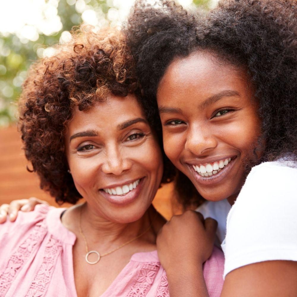 Smiling woman in pink blouse with arm around smiling young girl outdoors.