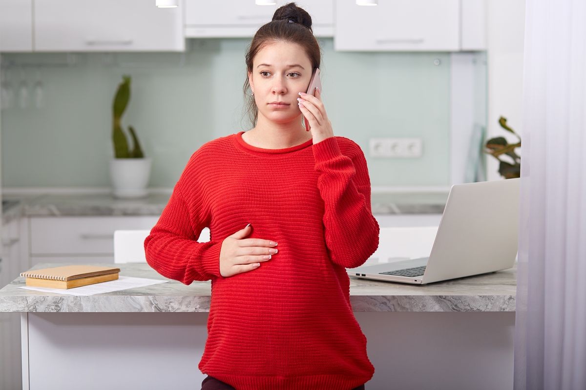 Image of pregant young woman keeps hand on tummy, dressed in red casual clothing, poses against kitchen interior, has telephone conversation, thoughtful expression, uses modern technologies.